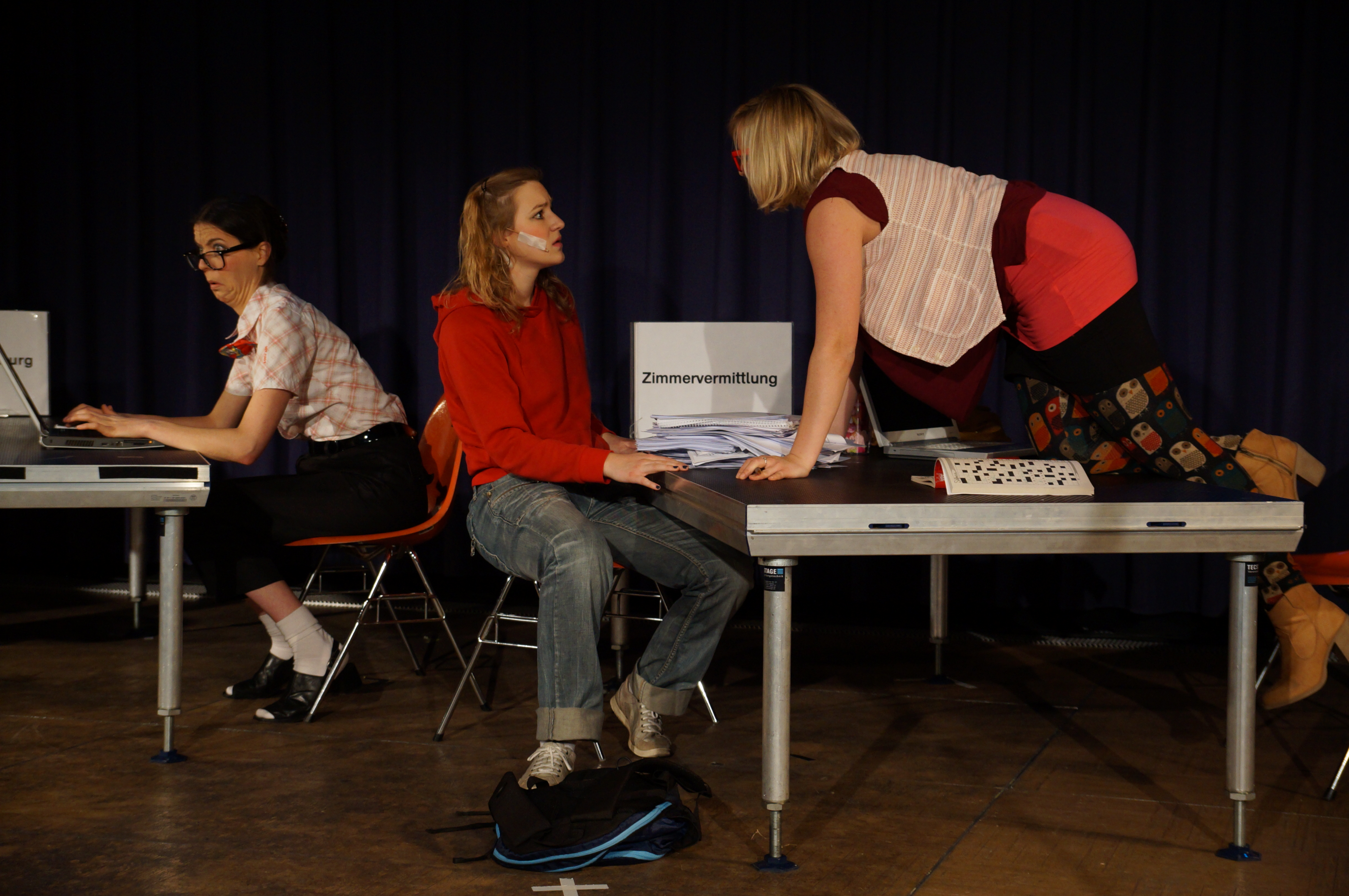 Eine Theateraufführung: auf der Bühne befinden sich zwei Tische und drei Schauspielerinnen: eine links sitzt am Computer. Rechts auf dem Bild, an dem zweiten Tisch befinden sich die zwei anderen Schauspielerinnen: eine sitzt auf einem Stuhl und schaut verwundert auf die dritte Frau, die in diesem Moment auf den Tisch steigt.